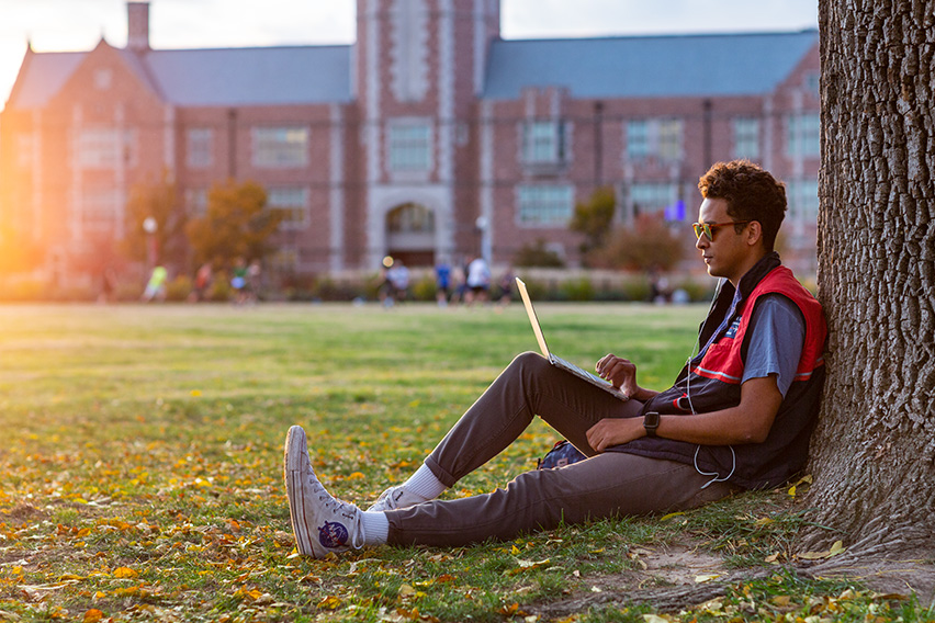 Student sits next to a tree on the WashU campus with his laptop open as the sun sets